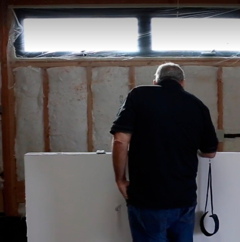 A person working in a partially constructed room with exposed insulation and framing, holding a white panel and measuring or inspecting.
