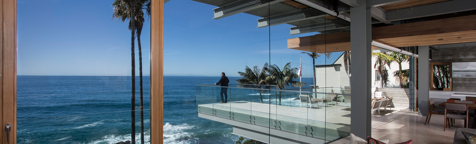 A stunning oceanfront view from a modern home with floor-to-ceiling windows, a glass balcony, and a person enjoying the scenery.