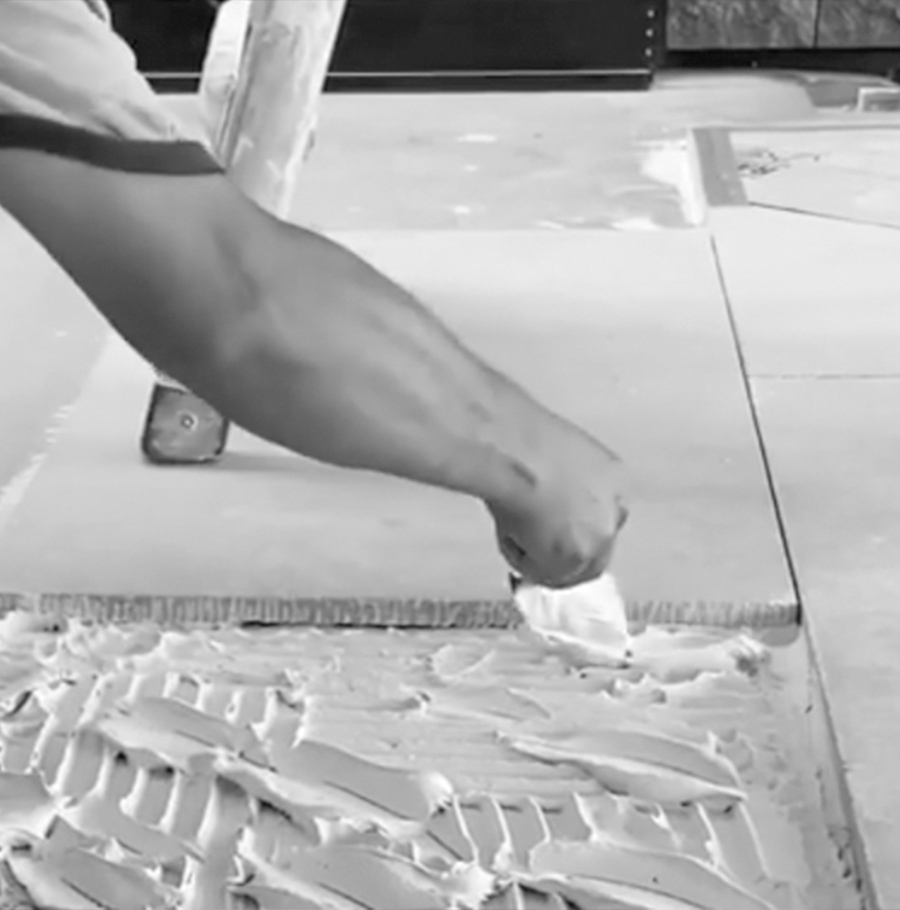 A black and white close-up of a person spreading adhesive with a trowel, preparing to lay tiles or stone on a flat surface.