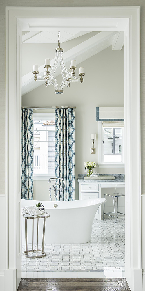 A luxurious bathroom with a freestanding white tub, chandelier, patterned curtains, white vanity, and elegant tile flooring.
