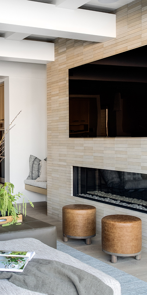 This image shows a modern living room with a beige brick-patterned wall, a large TV above a fireplace, leather stools, and a cozy seating area.
