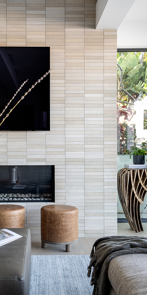 This image shows a modern living area with a beige brick-patterned wall, a TV, a fireplace, leather stools, and a decorative wooden table.