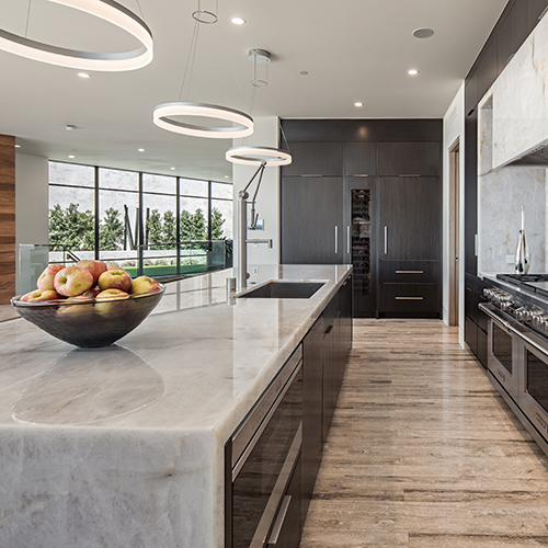 A sleek, modern kitchen with dark wood cabinets, a large marble island, contemporary pendant lights, and a bowl of apples on the counter.