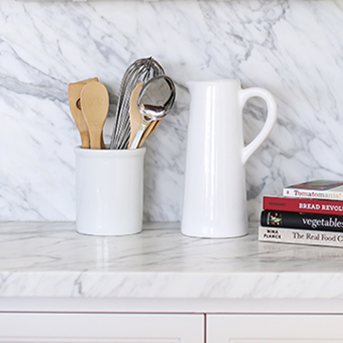 A kitchen countertop with a marble backsplash, white utensil holder with wooden spoons and a whisk, a white pitcher, and cookbooks.