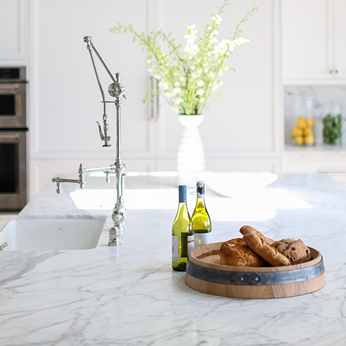 A bright kitchen with a marble countertop, chrome faucet, a vase with flowers, a wooden tray with bread, and two bottles of wine.