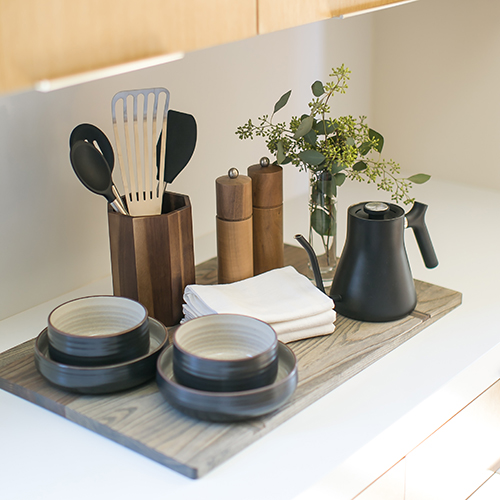 A kitchen counter with wooden utensil holders, black dishes, a black kettle, salt and pepper grinders, a vase with greenery, and folded napkins.