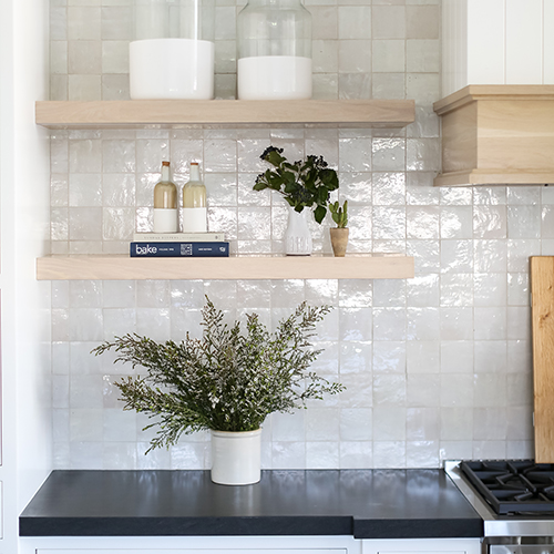 A kitchen with a glossy tile backsplash, light wood floating shelves, various decor items, and a potted plant on a dark countertop.