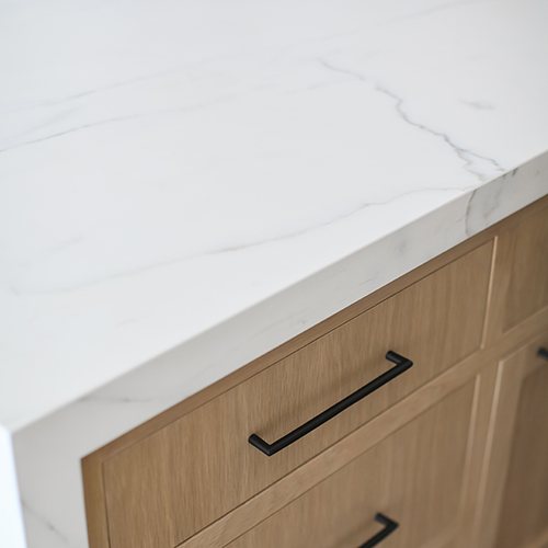 A close-up of a kitchen counter featuring a white marble countertop with subtle veining and light wood cabinets with black handles.
