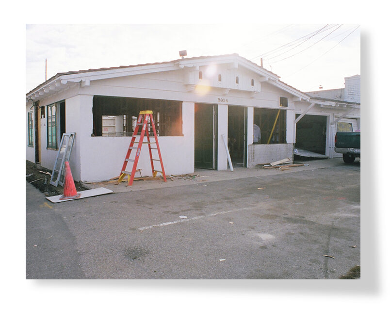 This image shows a single-story building under renovation, with large openings for new windows or doors, a red ladder, and construction materials scattered around.